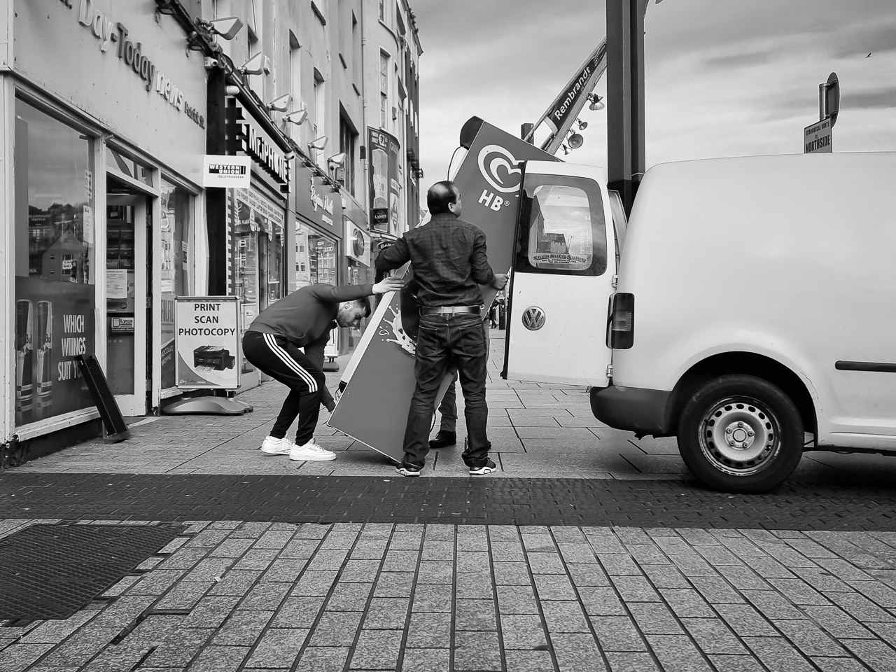 An ice cream fridge is loaded into a van from a shop on Patrick's Street, Cork. Two men hold the mid section while another man bends down to lift it up. The end of a van and it's doors are visible on the right.