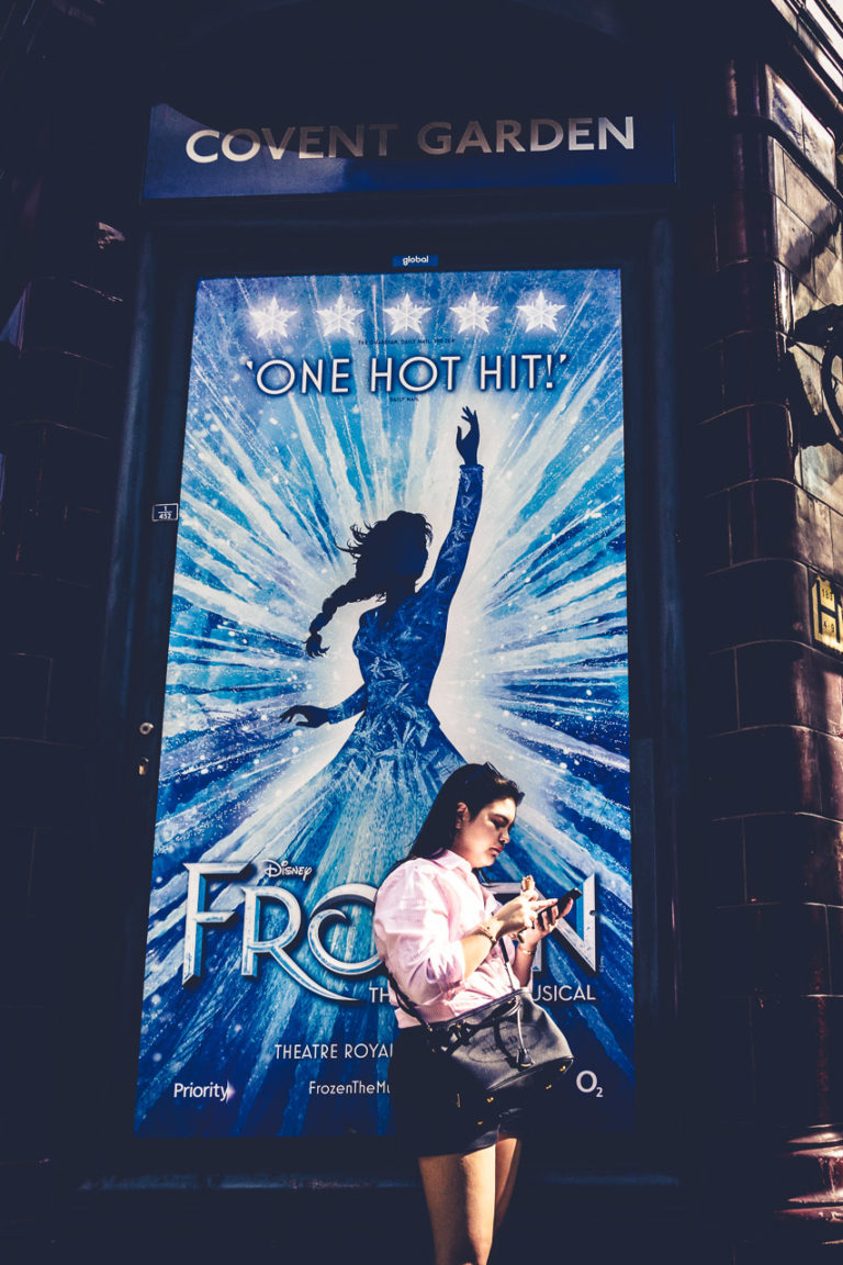A woman checks her phone in front of the Covent Garden Underground station, with a huge poster for Frozen, the musical, behind her.