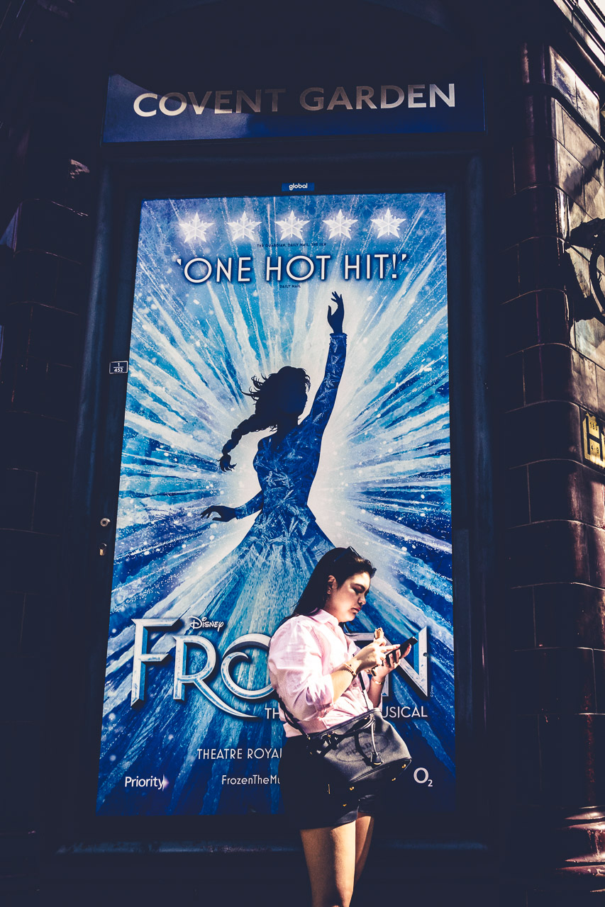 A woman checks her phone in front of the Covent Garden Underground station, with a huge poster for Frozen, the musical, behind her.