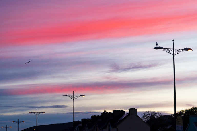 High clouds are lit by the sun after it has set. They're a vivid red thanks to the sun shining through the atmosphere. Street lights and the tops of houses are visible too.
