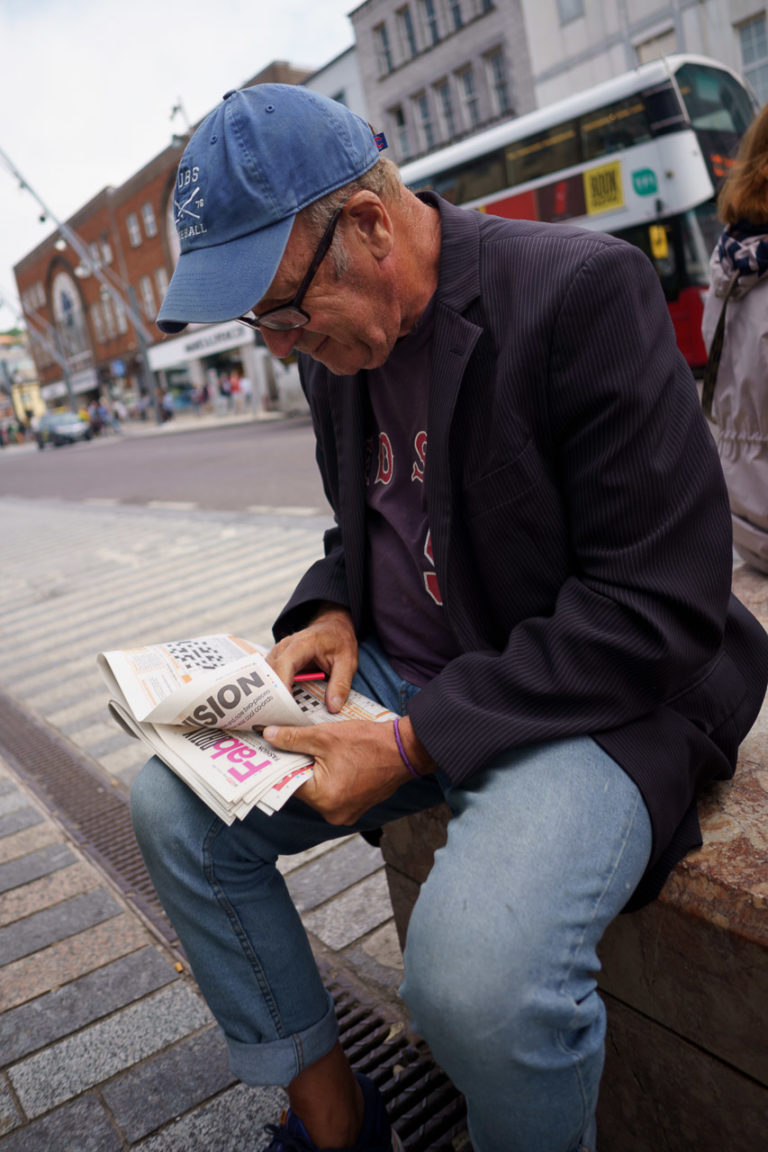 A man reading the Sun newspaper on the street. He wears a baseball cap, and is probably doing the crossword as he has a pencil in his hand. The word "Fab" is seen on an inside page of the paper.
