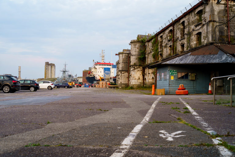The old, abandoned buildings behind the Customs House in Cork City. Cars are parked on the quay, a walkway is underfoot and boats are in the background.