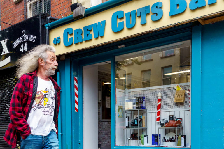 A man with a huge head of flowing hair walks past a barber's shop. He is wearing a red/black check jacket, a Looney Tunes tshirt and jeans