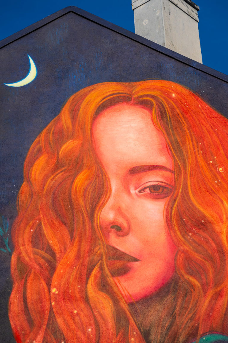 A detail from a mural showing a red haired woman. Her hair covers half her face. The Moon is visible in the background of the mural, while the top of the house forms an upside down V and a chimney sits on top.