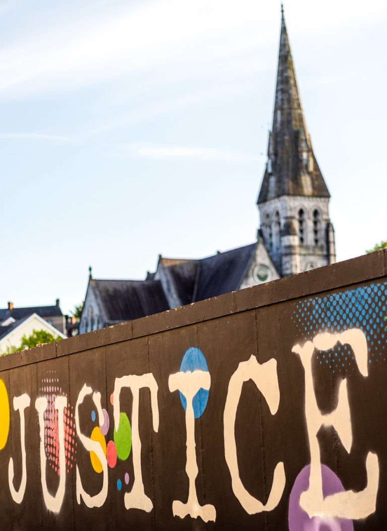 The word JUSTICE is painted on the hoarding around a derelict site on Sullivan's Quay. The spire of a deconsecrated church in the background.