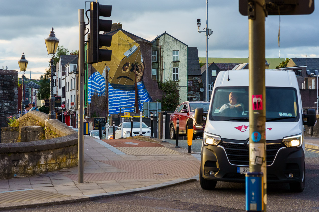 Car traffic and a white van crossed a bridge in front of me, going through a junction with traffic lights, while the "What is Home?" mural on South Main Street is visible in the background.