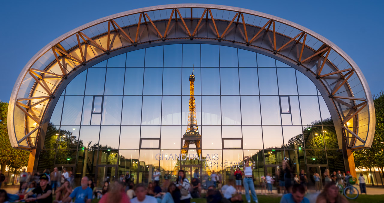 The Eiffel Tower reflected in the windows of the Grand Palais Éphémère in Paris. The sky is clear and sun has set creating a yellow glow on the horizon. People are seen in the foreground watching the tower.