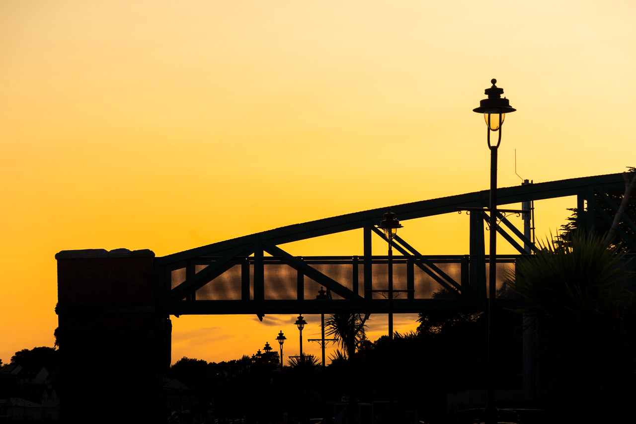 The footbridge over the railway line into Cobh captured in silhouette against a yellow sky cast by a setting sun.