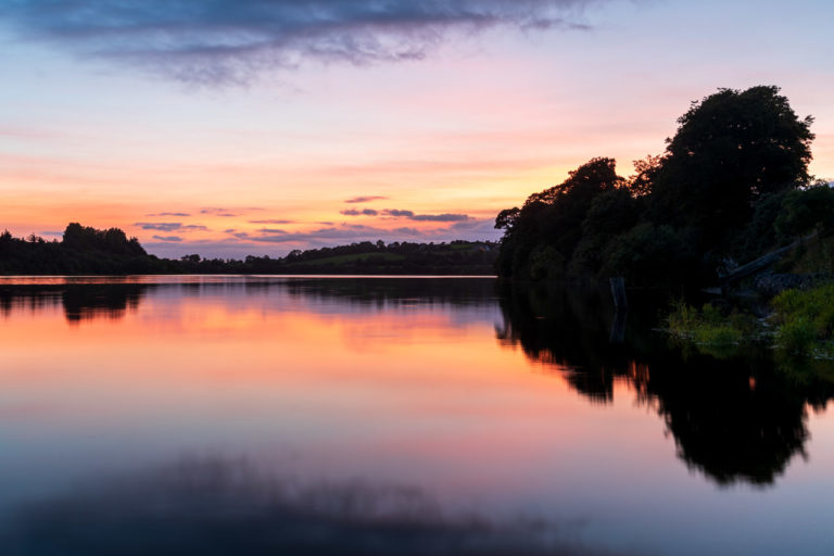 The orange, red and yellow glow in the sky after the sun has set is caused by the sun shining on the underside of high altitude cloud. It's reflected in the water and the land is in shadow.