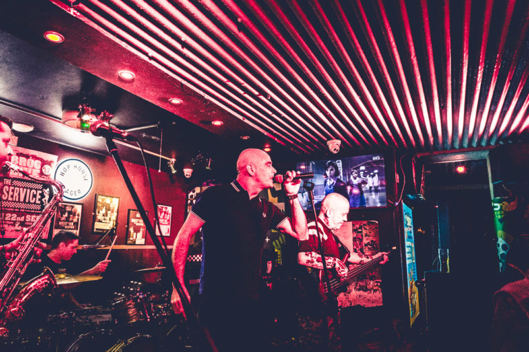 Members of the band, The Service, play on stage at An Brog. A singer, bass guitarist, Saxophone player and drummer are visible.