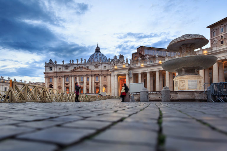 A woman takes the photo of another woman who leans against a small pillar near a fountain in St. Peter's Square, in the Vatican.