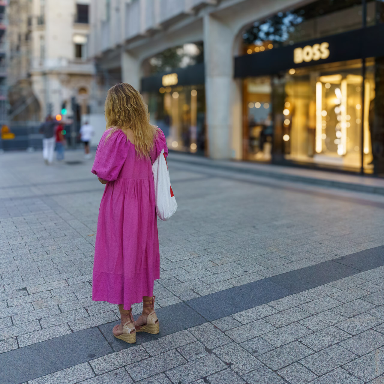 A woman in a pink dress stands on the street, in front of a BOSS store.