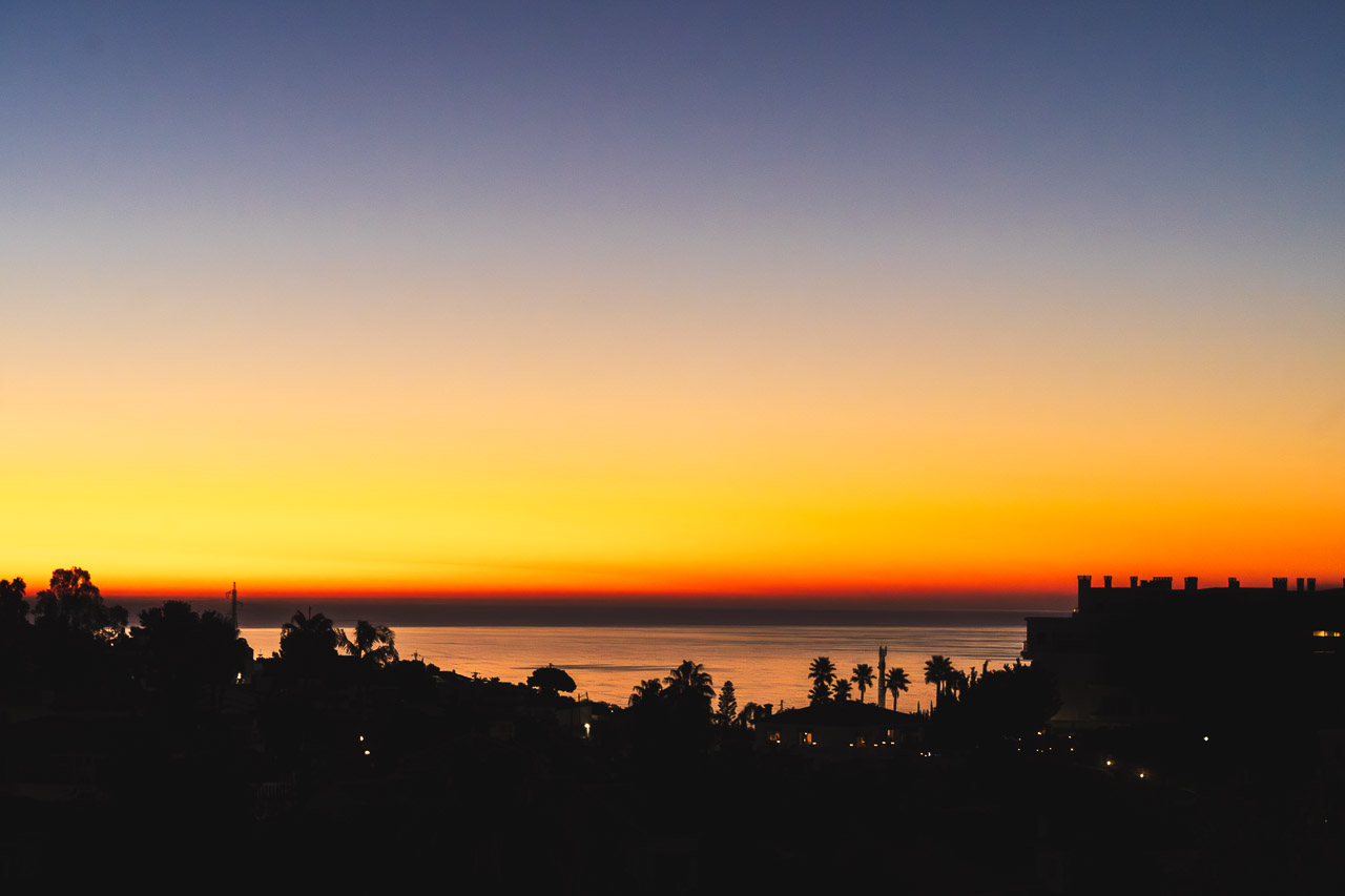 The glow of the sun before it rises above the horizon is silhouetted by houses and trees.