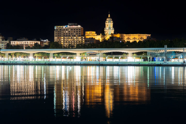 The Palmeral de Las Sorpresas, a promenade on Malaga Harbour is all lit up at night, with Malaga Cathedral in the background.