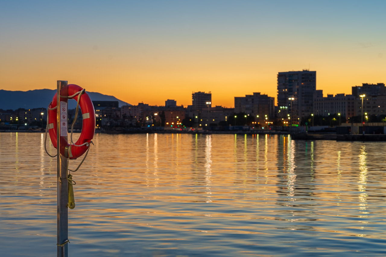 A lifebuoy on a pole with the water of Malaga Harbour behind it just after the sun has set.