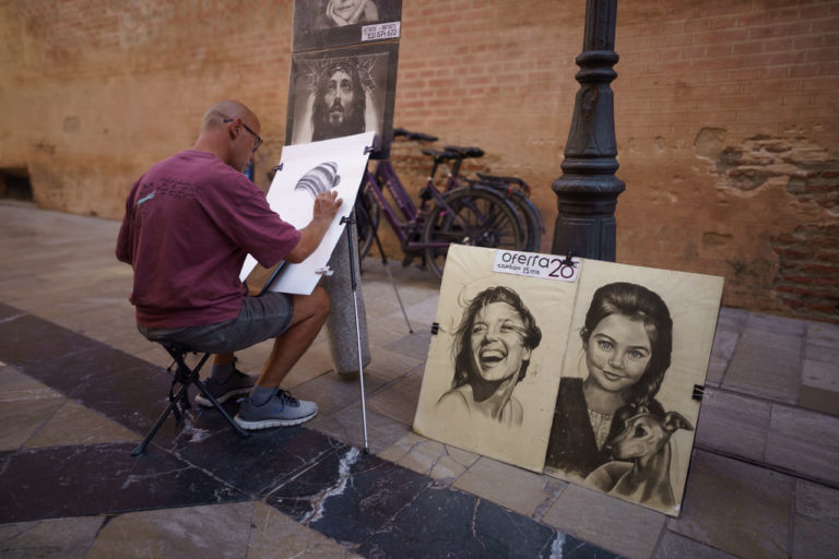 A street artists in Malaga works on a portrait. Other portraits sit on the path near his feet.