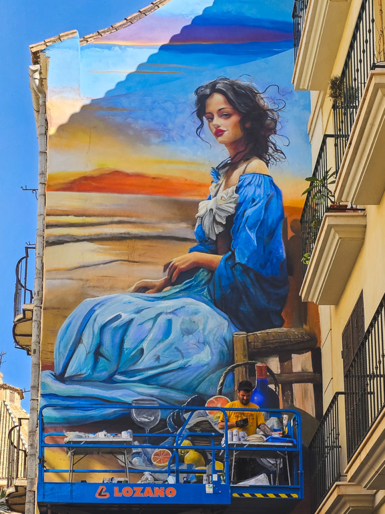 A mural of a woman in a blue dress. A man on a raised platform checks his phone and takes a break from painting.
