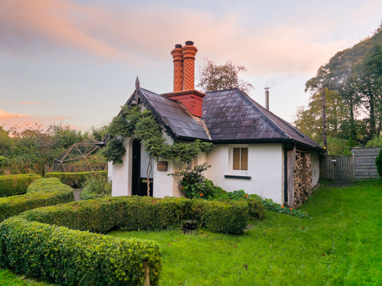 A small cottage surrounded by a neat lawn and hedges with trees in the background.