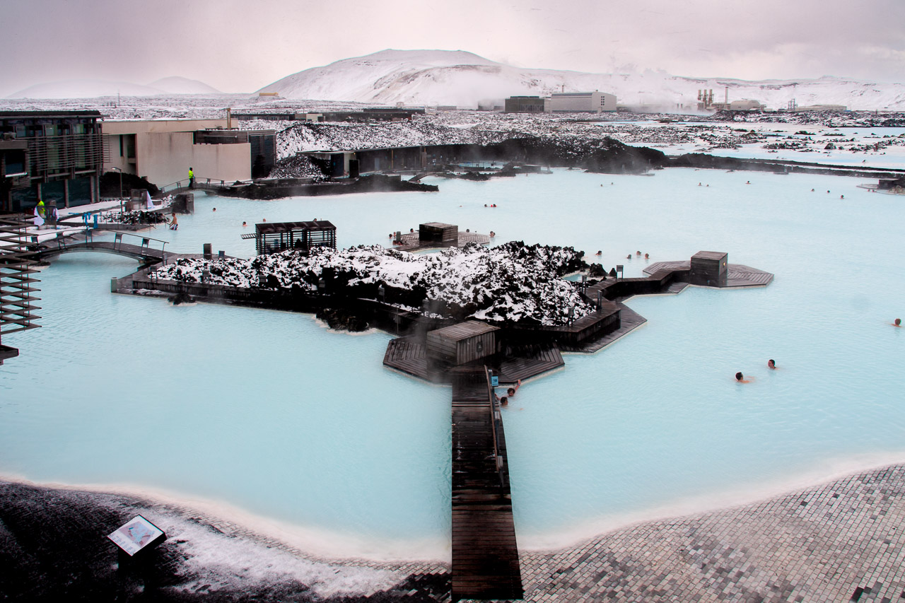 The blue waters of the Blue Lagoon in Iceland. People are swimming in it, while the surrounding landscape is covered in snow. The Svartsengi Geothermal Power Plant can be seen in the background.