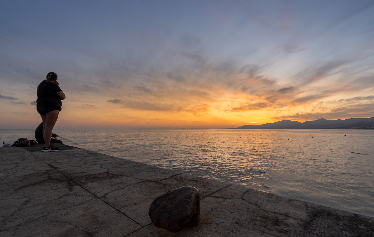 A man fishing off a quay in Puerto del Carmen, Lanzarote. His wife stands next to him and the sun sets over the water he's fishing from.