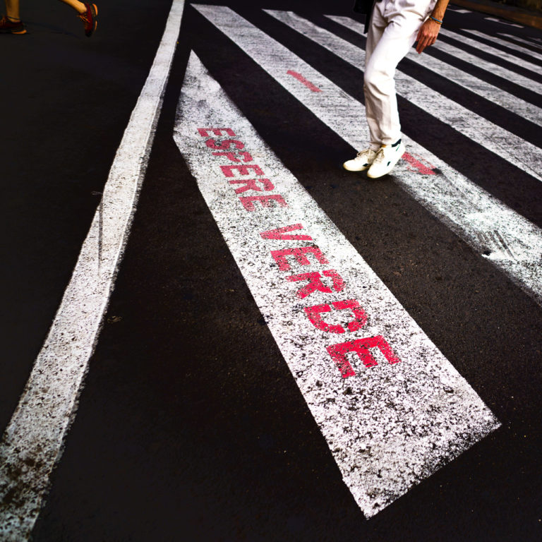 A person in white pants and runners walks across a street at a pedestrian lights. The white painted bars on the street are visible along with the words "Espere Verde". Another person has already crossed the street and their legs are seen departing the scene
