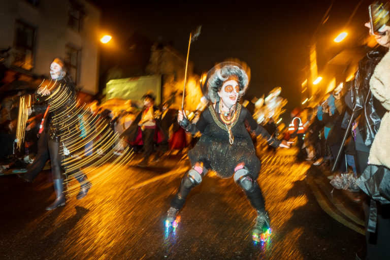 A blurry shot of a woman on roller skates taking part in the parade. She's got wonderful makeup and dressed in dark clothes, while nearby another woman swings lights. Behind them are others in the parade.