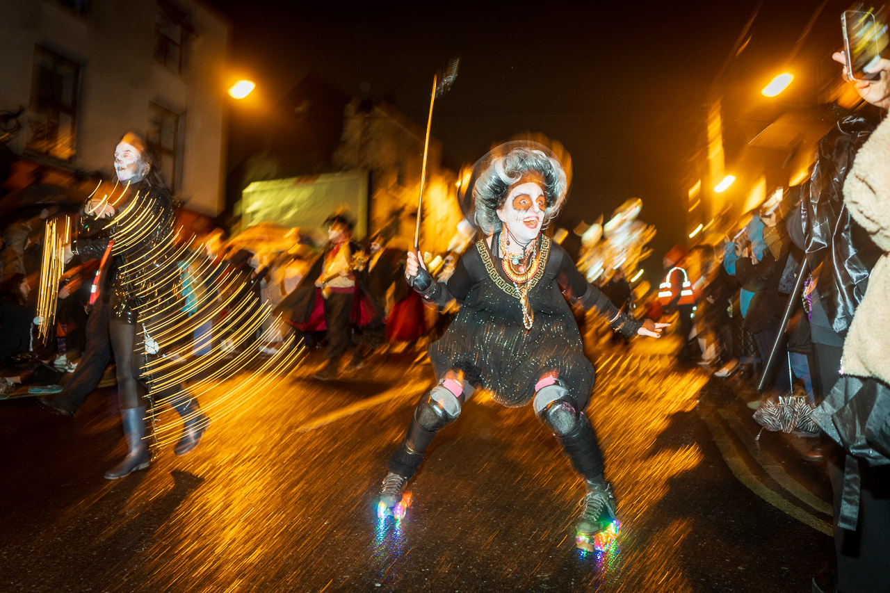 A blurry shot of a woman on roller skates taking part in the parade. She's got wonderful makeup and dressed in dark clothes, while nearby another woman swings lights. Behind them are others in the parade.