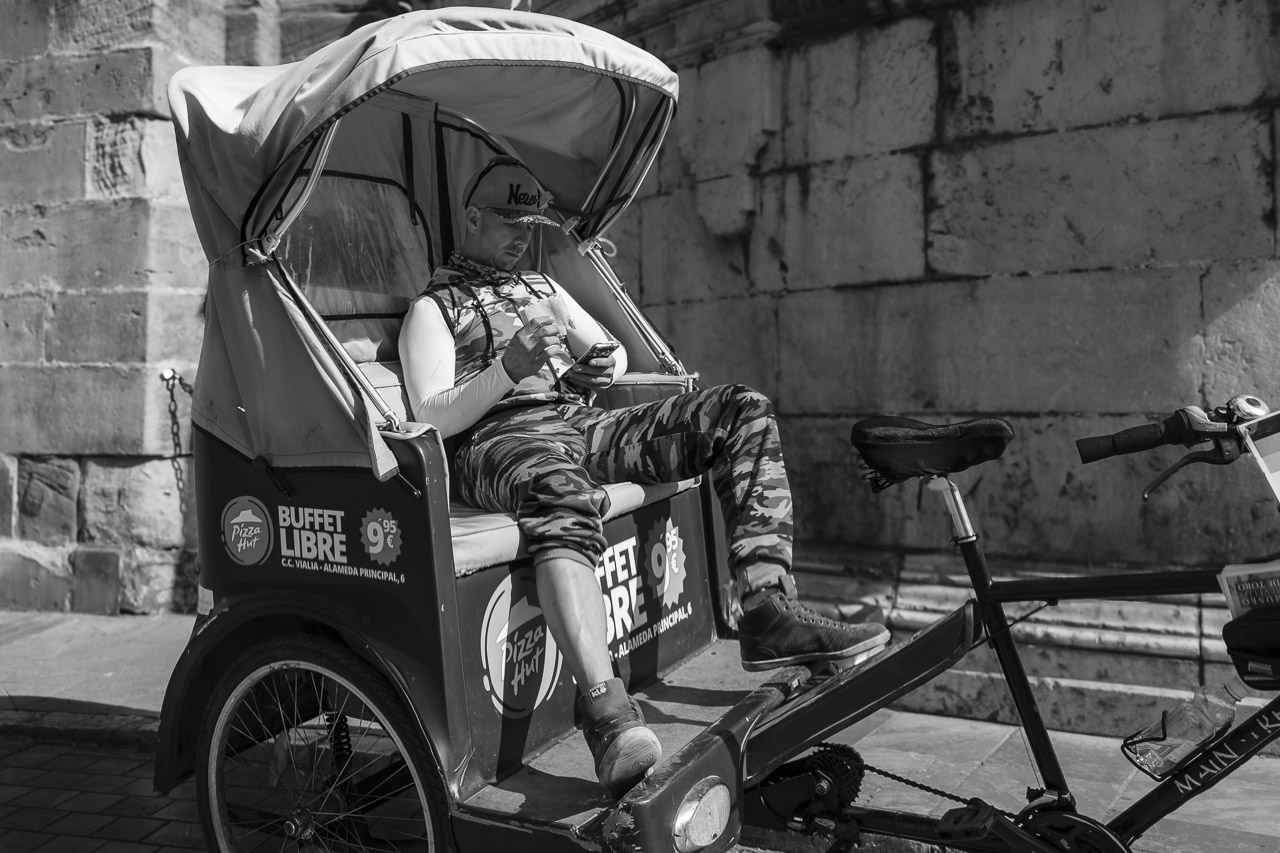 A man sitting in the back of an open tricycle, waiting for passengers. He appears to be relaxing and enjoying his time as he looks at his phone while seated.
