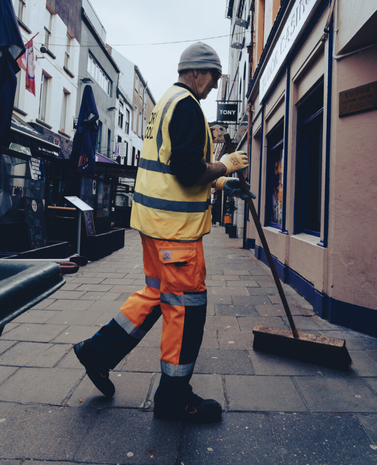 A man uses a large brush to sweep the stret. He is wearing reflective gear, gloves and a woollen beanie.