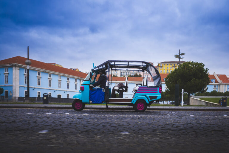 A blue tuk tuk driving along the road in front of a blue building, trees and in the far distance a yellow building. The sky has been coloured blue. Blue all over.