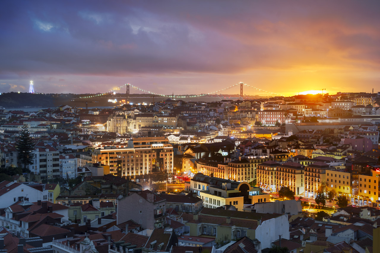 The city of Lisbon, with the statue of the Sanctuary of Christ the King and the suspension bridge, the Ponte 25 de Abril. The sun is setting.
