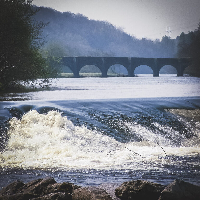 Water flows over a weird in the River Lee with trees visible on the bank, a bridge crossing in the background and poles visible in the far background.