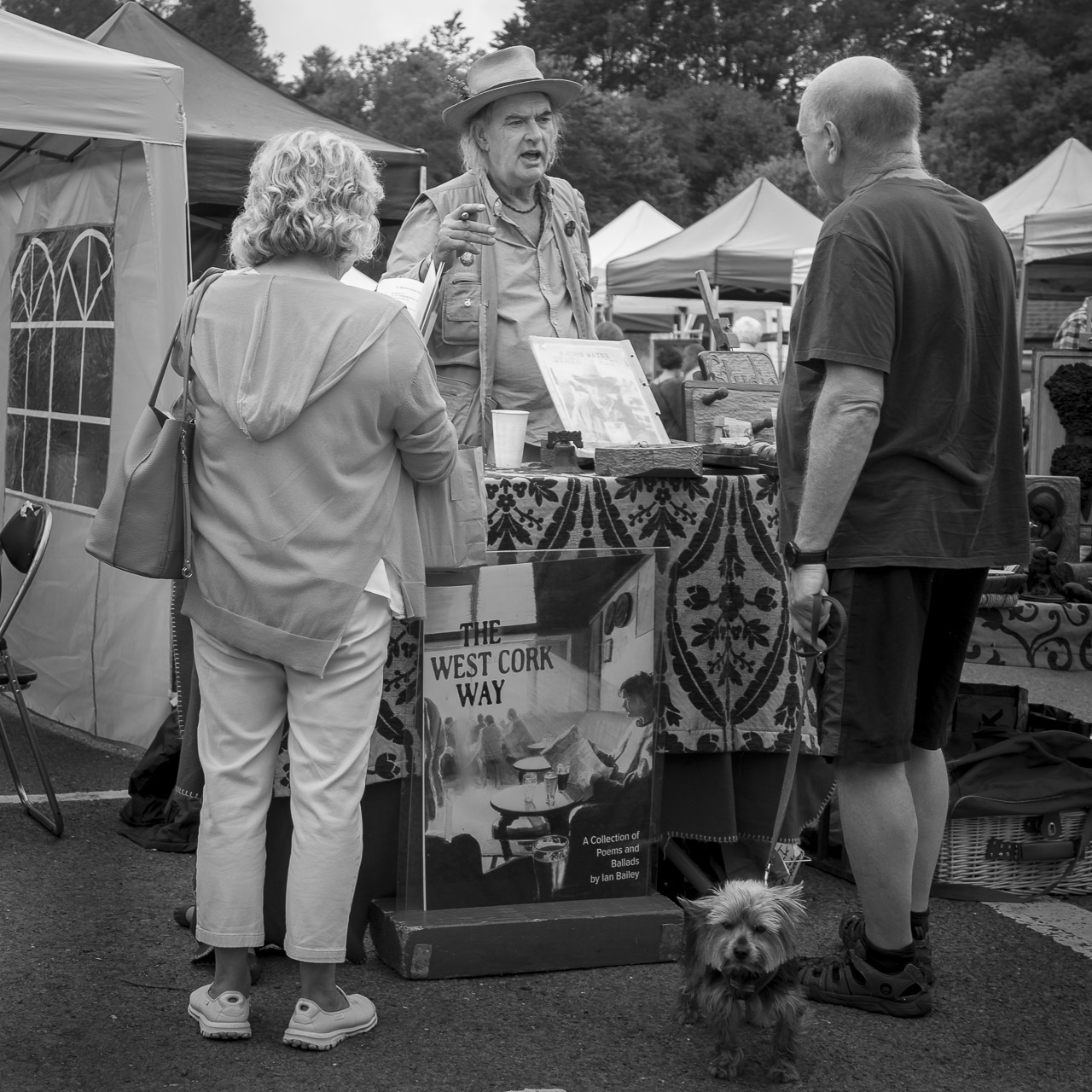 Ian Bailey at a stall in a market with the title of his book, The West Cork Way, on the front of it. He is talking to a man and a woman. The man is holding a lead with a small terrier dog.