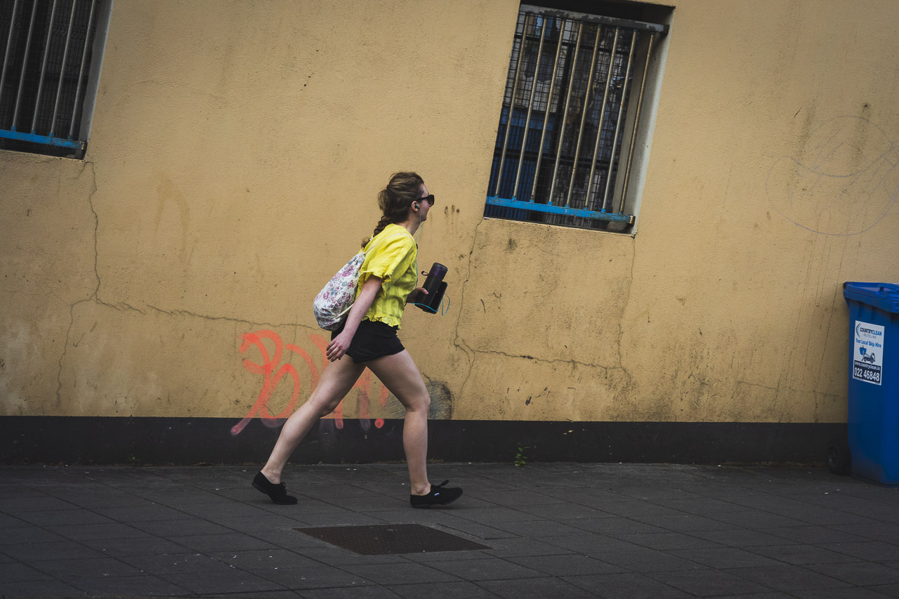 A woman in a yellow top, and dark shorts walks up a hill laid horizontal, with a yellow wall behind, and barred windows.