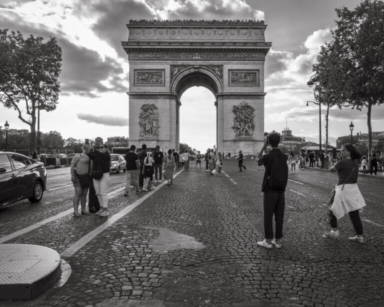 On a road leading to the Arc de Triomphe in Paris. Tourists line in the middle of the road, in relative safety from passing cars, but other tourists stop and take photos with their phones.