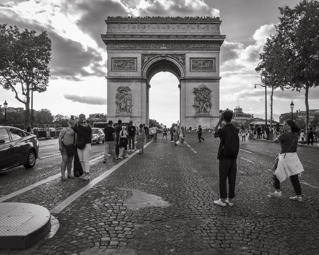 On a road leading to the Arc de Triomphe in Paris. Tourists line in the middle of the road, in relative safety from passing cars, but other tourists stop and take photos with their phones.
