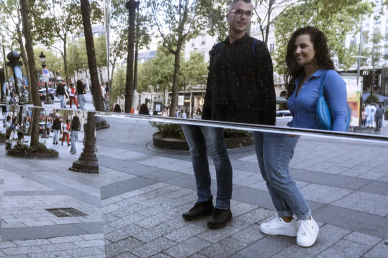 A couple look at themselves in the angled mirror facade of a building in Paris. Their legs are separated from their torsos by the reflection.