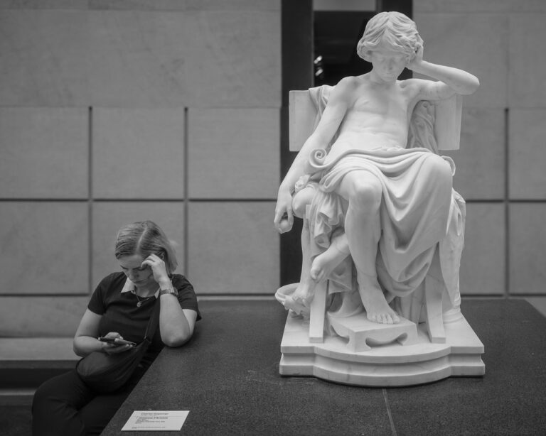 A woman sitting next to a statue unintentionally imitates the statue by resting her arm on the block the statue sits on.