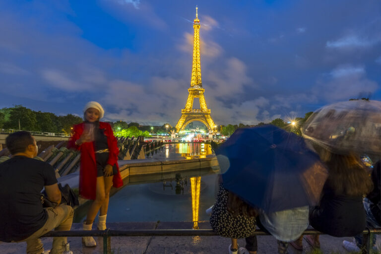A young woman poses for a selfie in front of the Eiffel Tower as it is all lit up. Other people are sitting down nearby. The Eiffel Tower is reflected in the waters of the Fontaine du Jardin du Trocadéro.