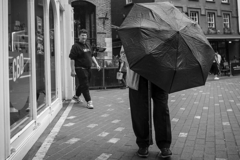 A man with a walking stick prepares to lower his umbrella as another man comes out of a nearby shop.