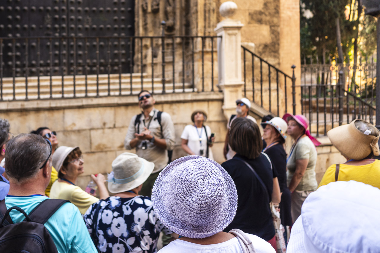 A tour group listens to their guide. Many of the people are wearing hats and one person who is one of the few people in focus in this photo is wearing a nice lilac hat that caught my attention.