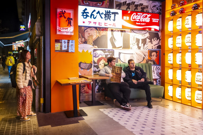 Two young men sitting on a couch, waiting to go into Buga Ramen. One holds a menu. A women looks in from the street, while signs hang on the wall, and one wall is made up of lanterns.