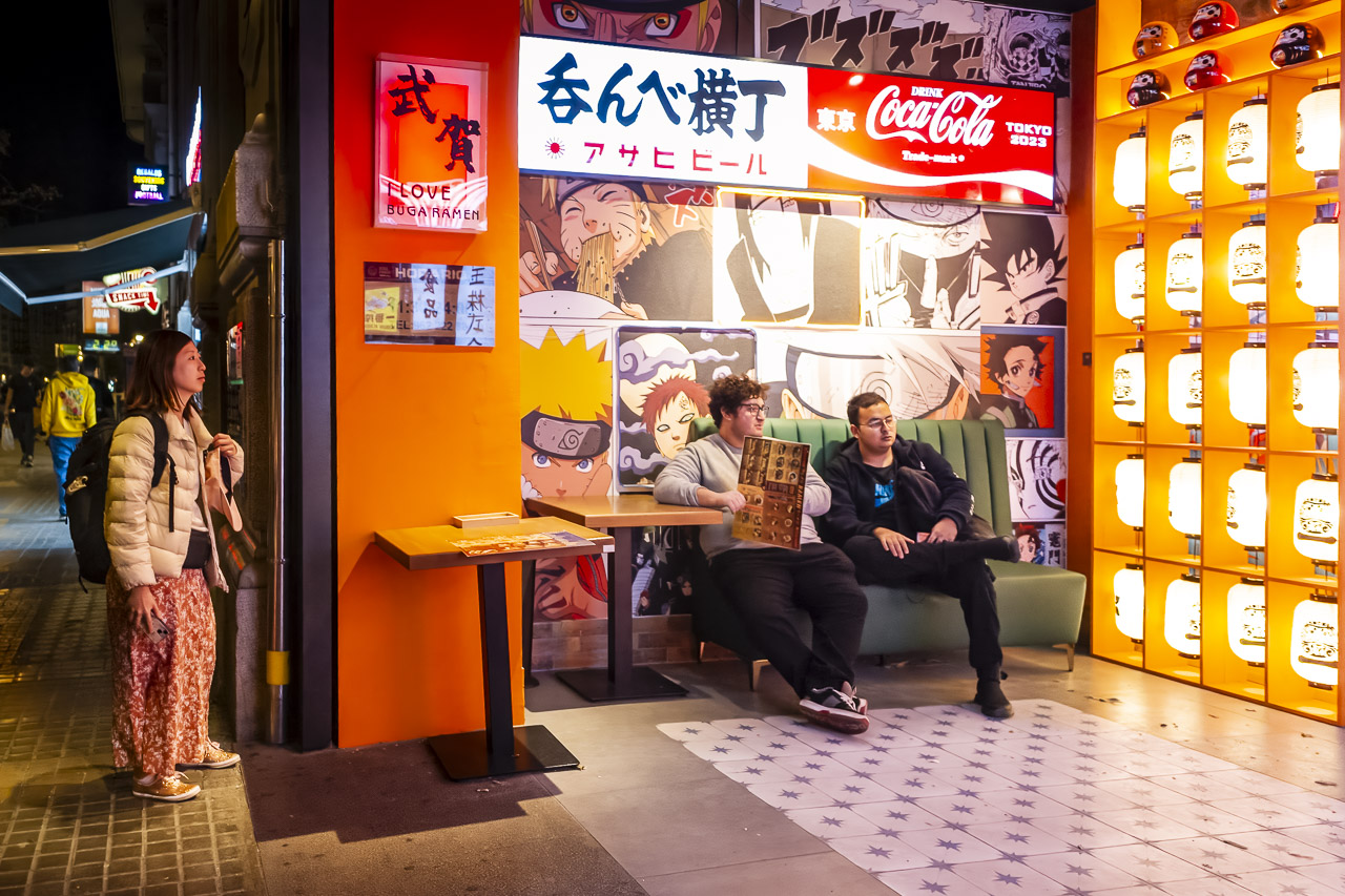 Two young men sitting on a couch, waiting to go into Buga Ramen. One holds a menu. A women looks in from the street, while signs hang on the wall, and one wall is made up of lanterns.