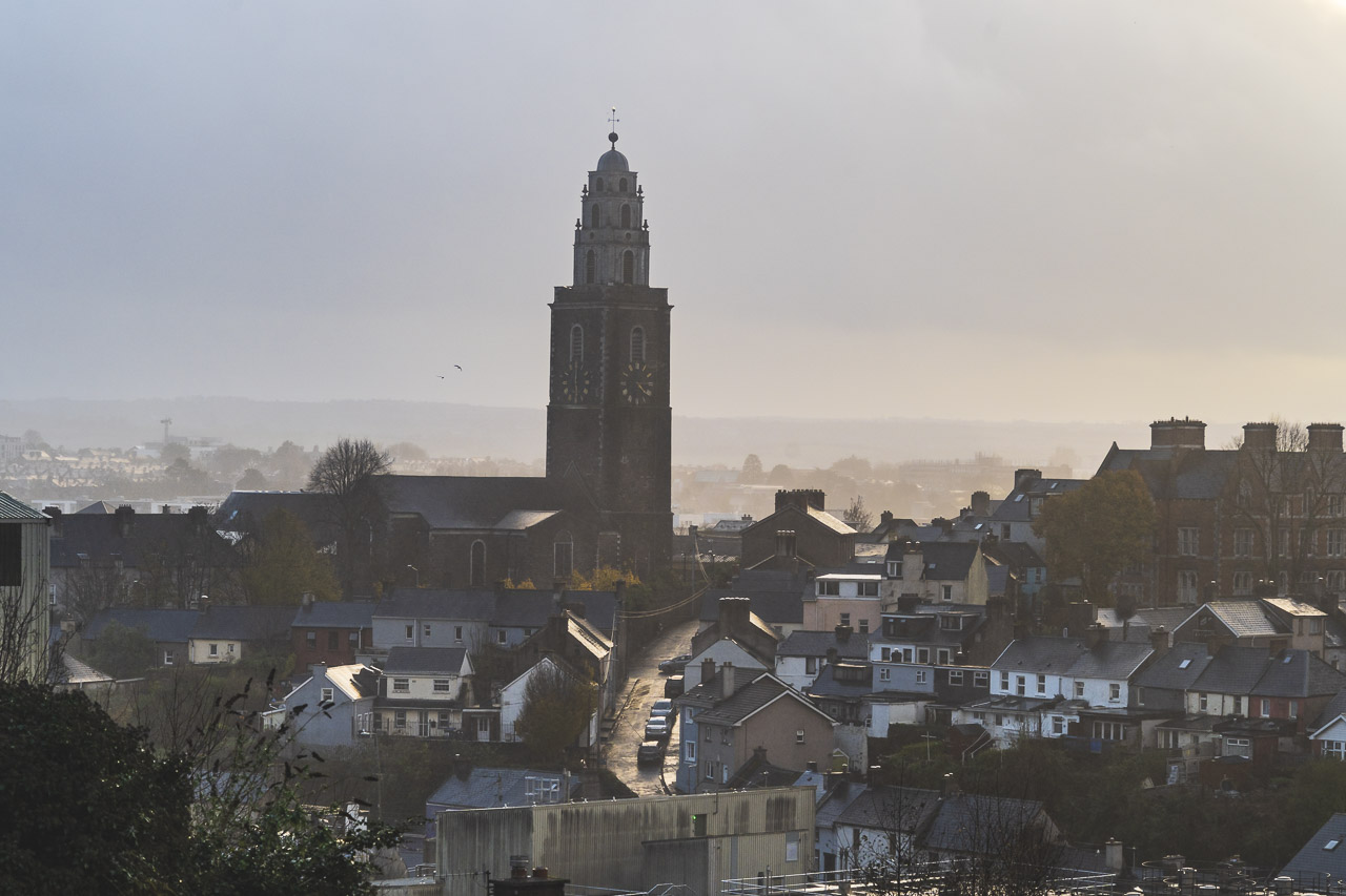 The spire of St. Anne's Church (Shandon Bells) rises above the surrounding houses and street next to it at sunset with cloudy skies overhead.