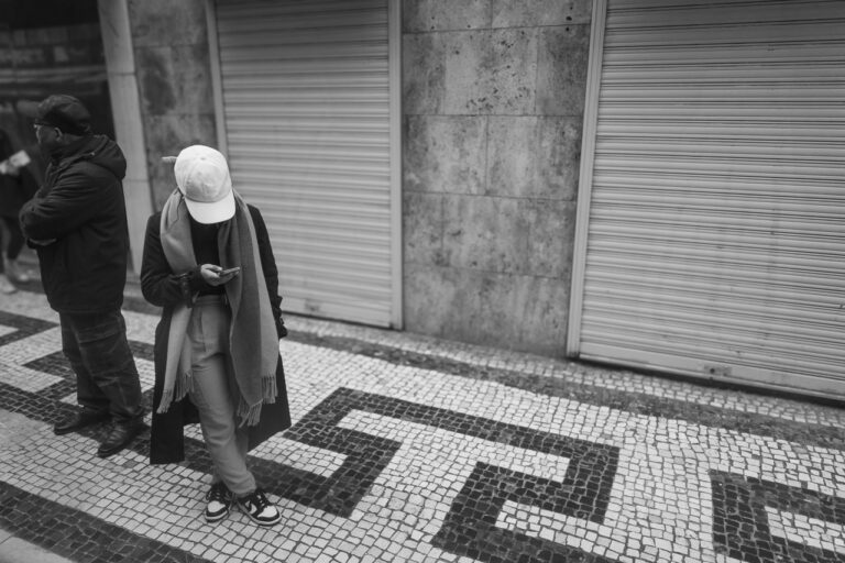 A person with their head down looking at a phone. They're wrapped up warmly against the (IMO) mild October weather in Lisbon. Another man next to them looks off in another direction. They're standing on a pavement made of tiny square tiles in a decorative pattern.