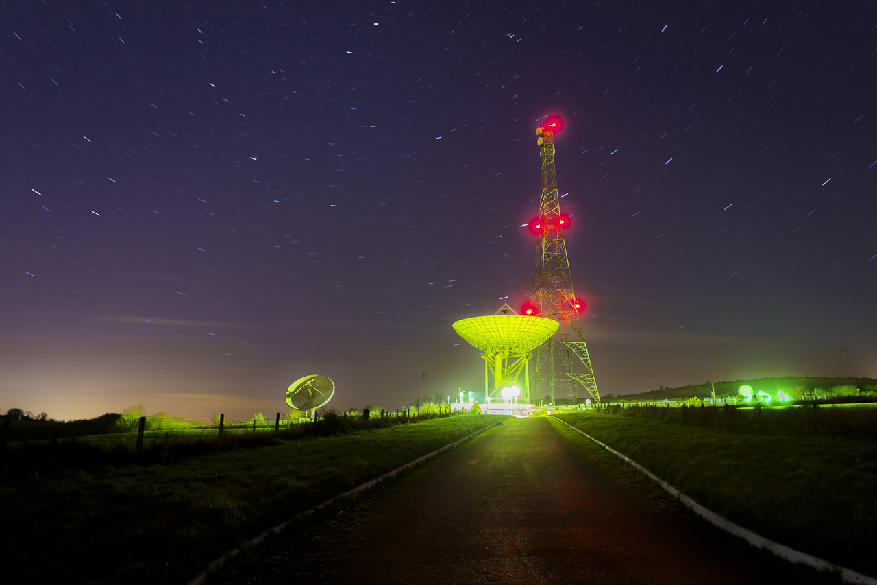 The National Space Centre at Elfordstown in Co. Cork is lit up at night. A large central dish is seen at the end of a road, some buildings underneath, a large radio tower next to it and a small dish visible to the left. Stars revolve in the sky in this long exposure shot.