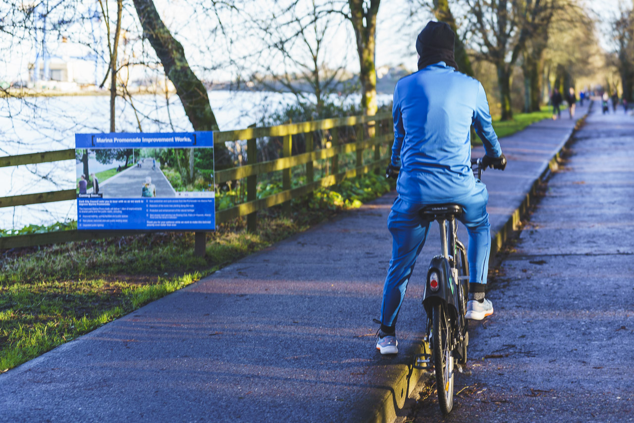 A cyclist reads a sign next to a road. The sign is blurred due to a narrow depth of field, but is about the Marina Promenade Improvement Works.
