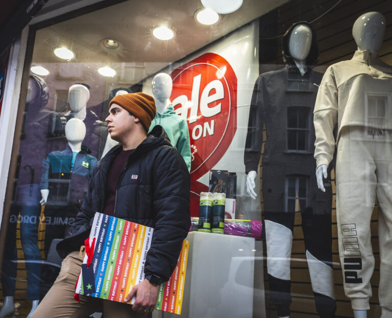 A man leans on a shop window, carrying a massive birthday card. Inside the shop, seven mannequins with faces heads watch out the window.