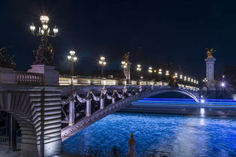 The Alexandre III Bridge in Paris pictured at night. A passing boat casts a blue light on the river.
