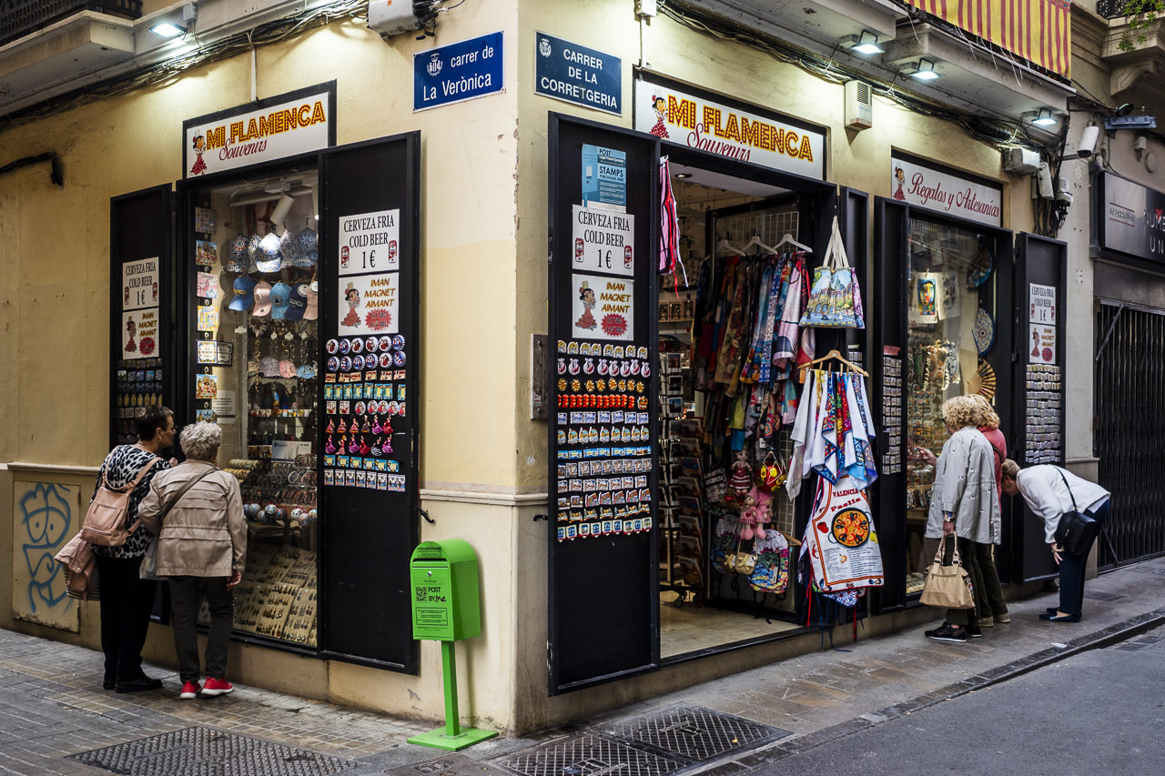 Five women look in a shop call MI FLAMENCA through two separate windows at the tourist knick knacks for sale. They're maybe unaware of each other as they are separated by a corner. A postbox sits at the corner, waiting for postcards home.